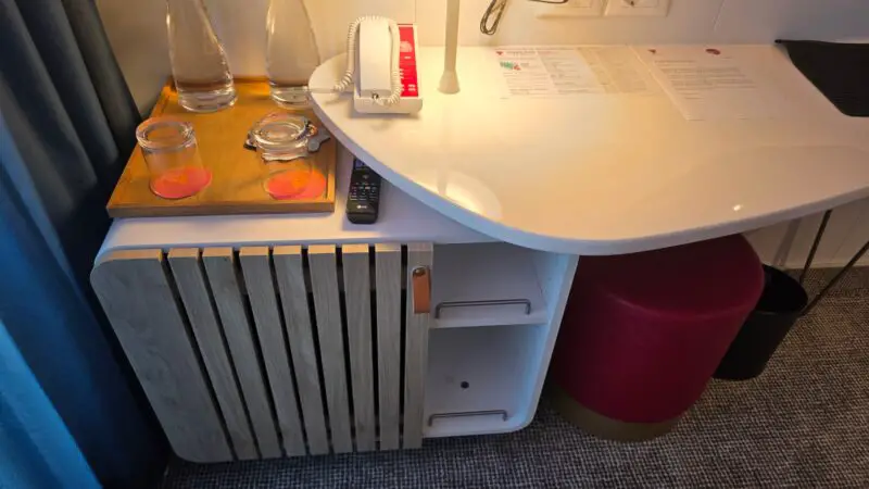 Modern cruise ship cabin with a slatted wood cabinet, topped with water carafes and glasses. Nearby is a curved white desk with a phone, outlets, and documents, above a red stool and carpeted floor.