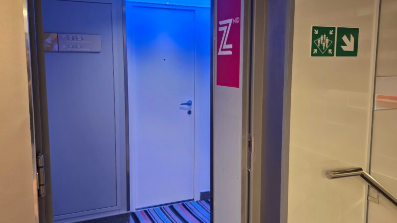 View into a Virgin Voyages cruise ship hallway with striped carpet and blue lighting. A door to a cabin is visible.