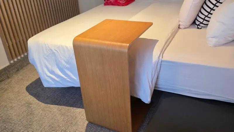 Virgin Voyages cruise ship cabin with a bed featuring white and striped pillows, a wooden bedside table, wall-mounted TV, and a folded red towel on a shelf.