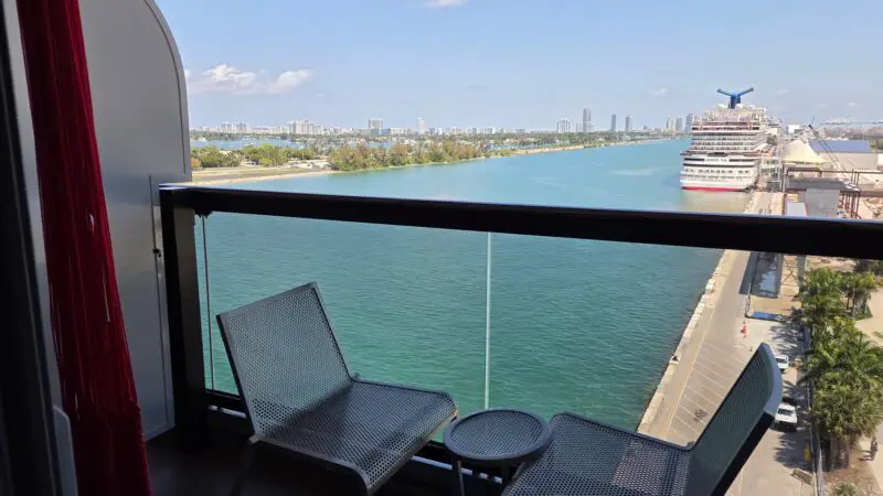 Balcony view from a Virgin Voyages cruise ship cabin, featuring two chairs, a small table, and a seaside port with another cruise ship in the background.