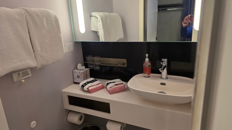Bathroom of a Virgin Voyages cruise ship cabin with a sink, mirror, towels, and toiletries.