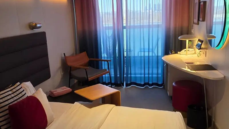 Virgin Voyages cruise ship cabin with a bed, accent pillows, a desk with a circular mirror, armchair, and balcony access through sheer, colorful curtains.