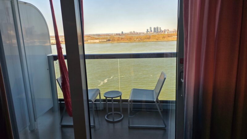 View from a Virgin Voyages cruise ship cabin showing a balcony with chairs and a table overlooking the water and distant city skyline.