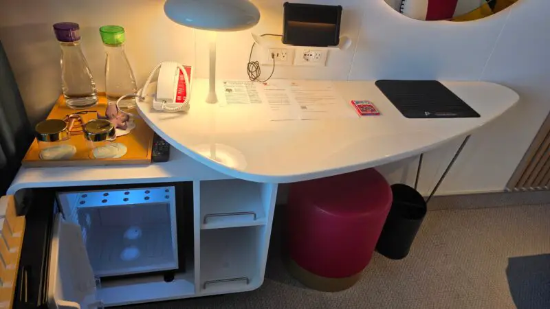 Virgin Voyages cruise ship cabin with a modern desk setup, featuring water bottles, a lamp, telephone, and an open mini-fridge below.