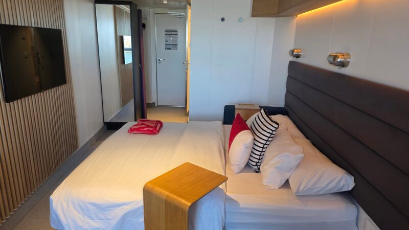 Virgin Voyages cruise ship cabin with modern design featuring a bed with striped and red pillows, wall-mounted lights, a wooden accent wall, and a TV.