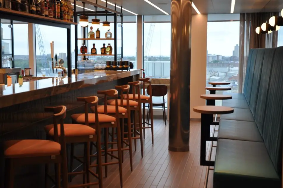 Bar area on a Virgin Voyages cruise ship with a marble counter and wooden stools, city view through windows
