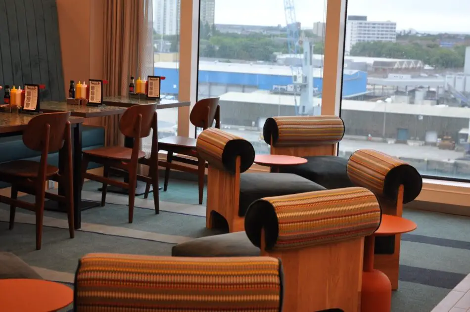 Cruise ship lounge area with striped upholstered chairs, orange side tables, and a large window overlooking the port