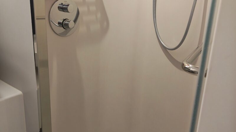 Shower area in a Virgin Voyages cruise ship cabin, featuring body wash dispensers, a showerhead, and a shelf with a purple bath scrub.