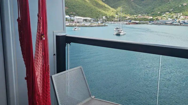 Balcony of a Virgin Voyages cruise ship cabin featuring metal chairs, a small table, and a red hammock with a scenic view of the sea and green mountains.