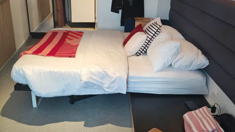 Virgin Voyages cruise ship cabin with a cozy bed, pillows, and a blanket, featuring stylish wood and upholstered accents.