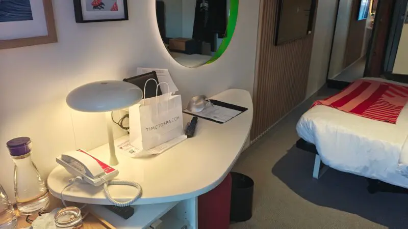 Virgin Voyages cruise ship cabin with a desk featuring a lamp, phone, and drinks, a bed with a red striped blanket, and wall-mounted TV.