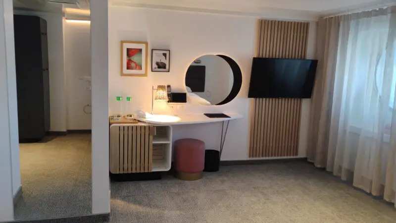 Modern Virgin Voyages cruise ship cabin with a round mirror, desk, wall-mounted TV, and window with sheer curtains.