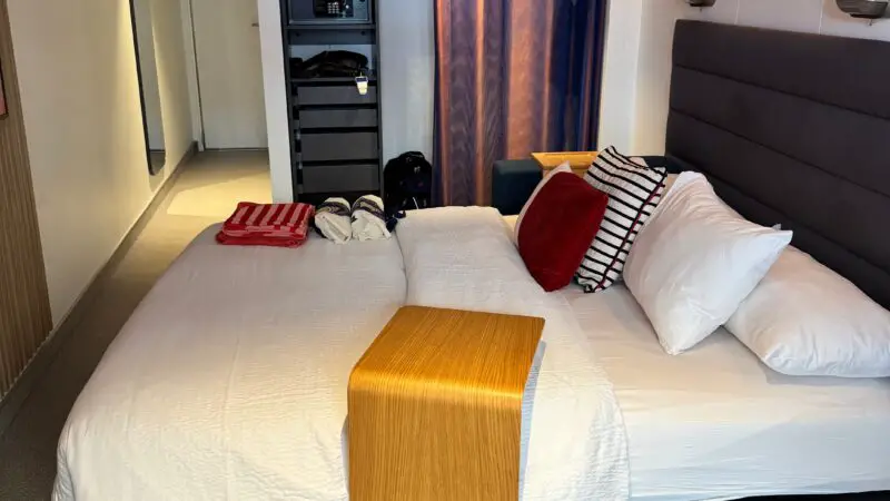 Virgin Voyages cruise ship cabin featuring a bed with white linens, wooden side table, and a storage area with a safe. The cabin includes a curtain separating the sleeping area from the closet space.