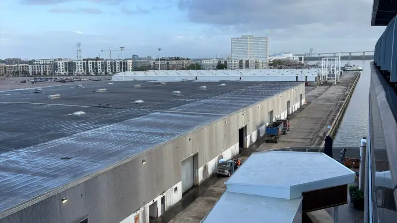View from a Virgin Voyages cruise ship balcony showing an industrial port with warehouses, vehicles, and a clear sky.