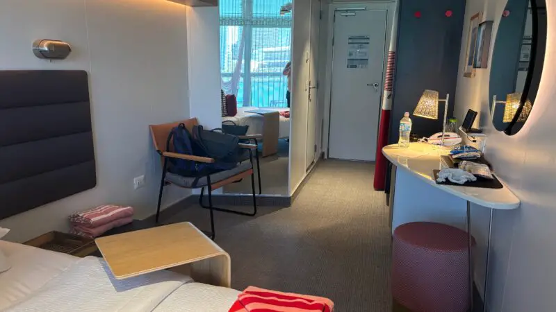 Virgin Voyages cruise ship cabin interior with a bed, chair, desk, and small table. A balcony with a hammock is visible through glass doors.