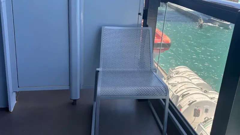 A Virgin Voyages cruise ship cabin balcony with a metal chair and table overlooking the water and city skyline.