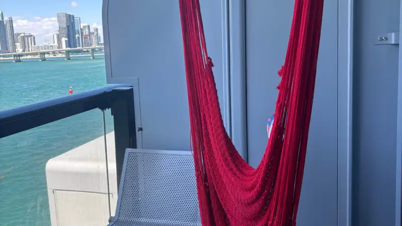 Balcony of a Virgin Voyages cruise ship cabin with a red hammock, white chair, and small round table; ocean and city skyline in the background.