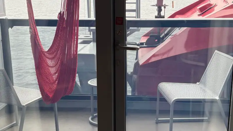 Virgin Voyages cruise ship cabin balcony with a red hammock, two white chairs, a small table, and an outside view of the ocean.