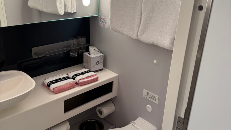 Modern bathroom in a Virgin Voyages cruise ship cabin, featuring a toilet, a wall-mounted mirror with a light, a countertop with a sink, towels, and a small waste bin.