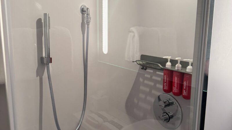 Shower area in a Virgin Voyages cruise ship cabin, featuring a handheld showerhead, modern fixtures, and red toiletries.