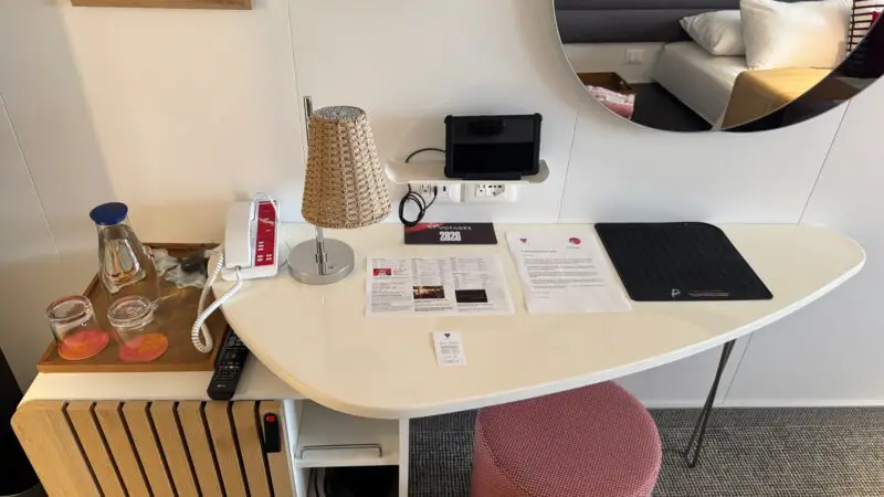 Virgin Voyages cruise ship cabin with a desk featuring a lamp, a phone, reading materials, and a tablet. A bed is partially visible reflected in a round mirror.