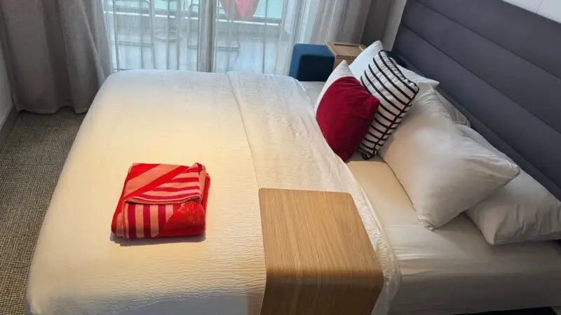 Virgin Voyages cruise ship cabin with a neatly made bed, colorful pillows, a folded striped towel, and a balcony with a hammock visible through glass doors.