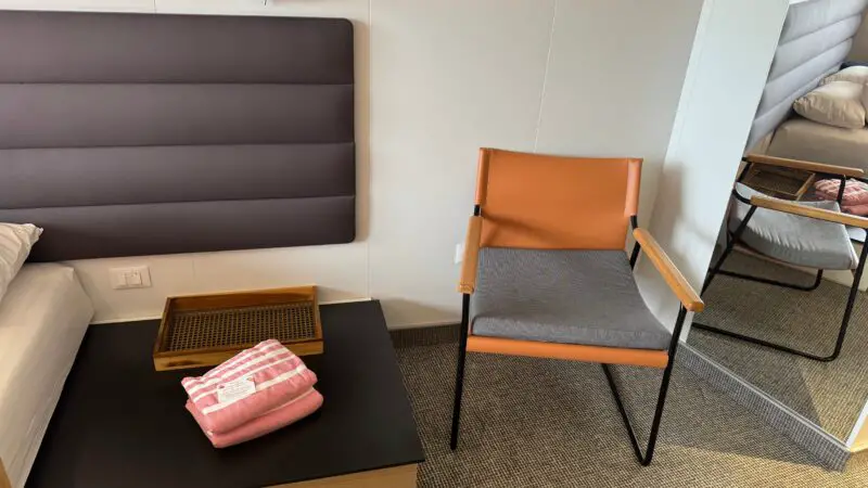 Virgin Voyages cruise ship cabin with a modern chair, bedside table, and neatly folded pink towels on the table.