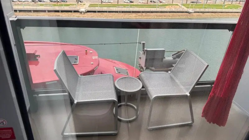 Balcony of a Virgin Voyages cruise ship cabin, featuring two modern chairs and a small round table. The glass barrier offers a view of the water and a distant shoreline lined with palm trees.
