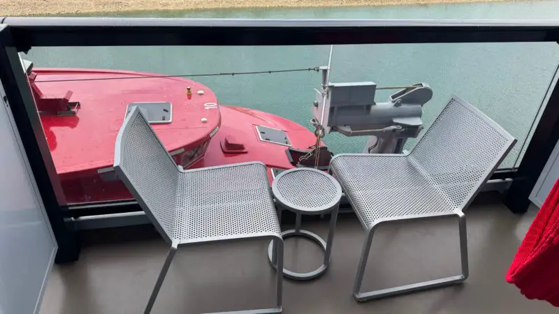 Balcony of a Virgin Voyages cruise ship cabin featuring two metal chairs and a small round table, overlooking the water and a red structure on the ship.