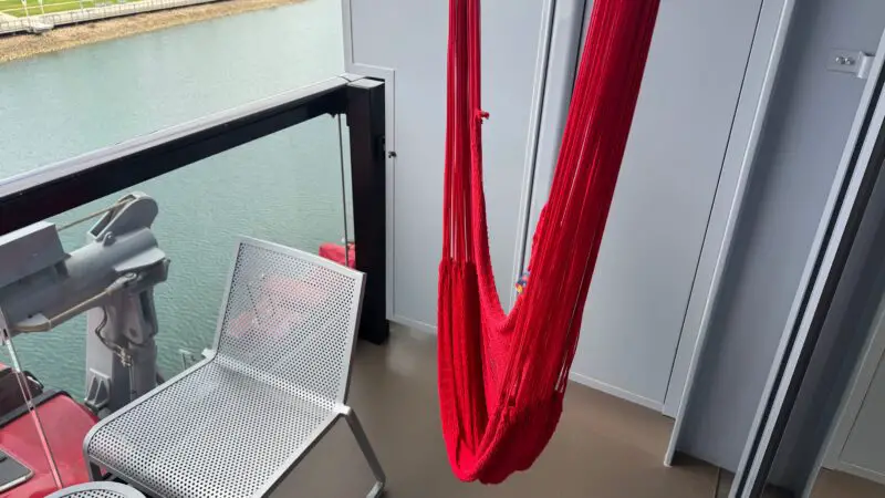 Balcony of a Virgin Voyages cruise ship cabin featuring a red hammock, metal chair, and small table overlooking the ocean.