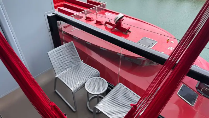 Virgin Voyages balcony cabin with a red hammock, two metal chairs, a small table, and a view of a red lifeboat and the sea.