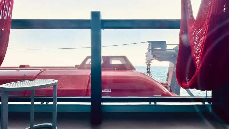View from a Virgin Voyages cruise ship cabin balcony, featuring an ocean view and a red hammock near a small white table, with part of a red lifeboat visible outside.
