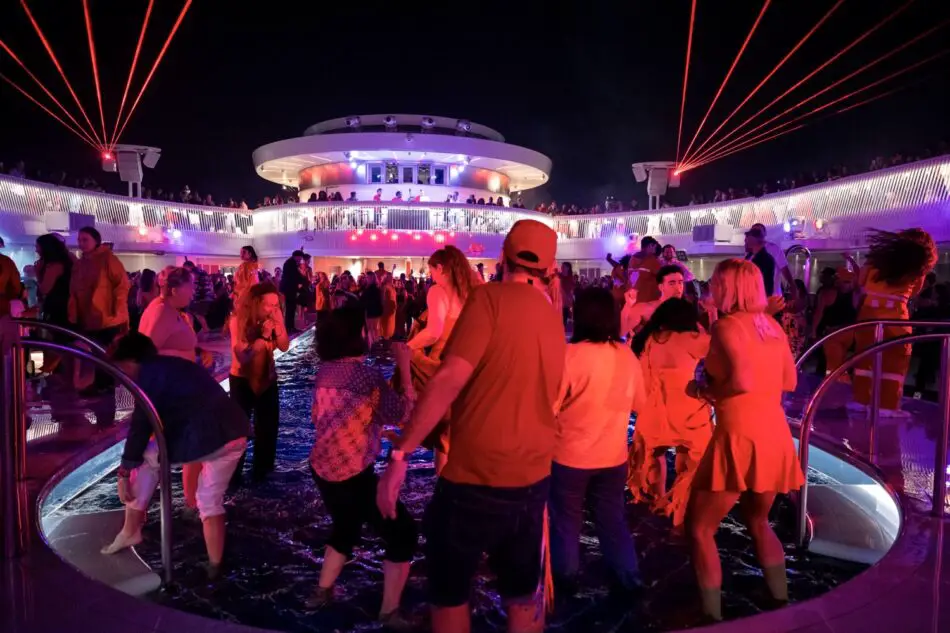 Crowd dancing around a lit pool on a Virgin Voyages cruise ship at night with red lasers
