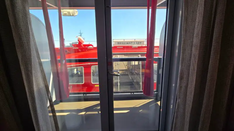 View from a Virgin Voyages stateroom balcony showing a red lifeboat and part of the ship's exterior labeled "Brilliant Lady", with blue sky in the background.