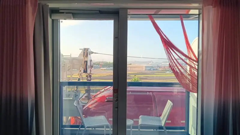 Virgin Voyages cruise ship cabin with a balcony view, featuring a red hammock, two chairs, and a small table outside sliding glass doors.