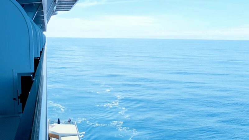 View from a Virgin Voyages cruise ship balcony, showing the ocean and a red lifeboat on the deck below.
