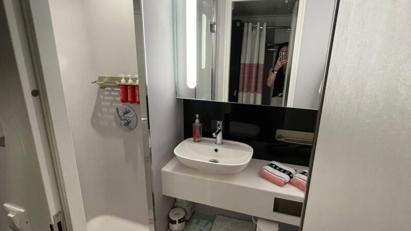 Bathroom in a Virgin Voyages cruise ship cabin featuring a shower, sink, large mirror, countertop with toiletries, and a toilet.