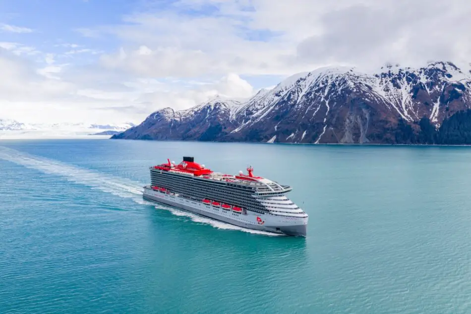 Virgin Voyages cruise ship with red upper decks sailing through turquoise water near snow-capped mountains