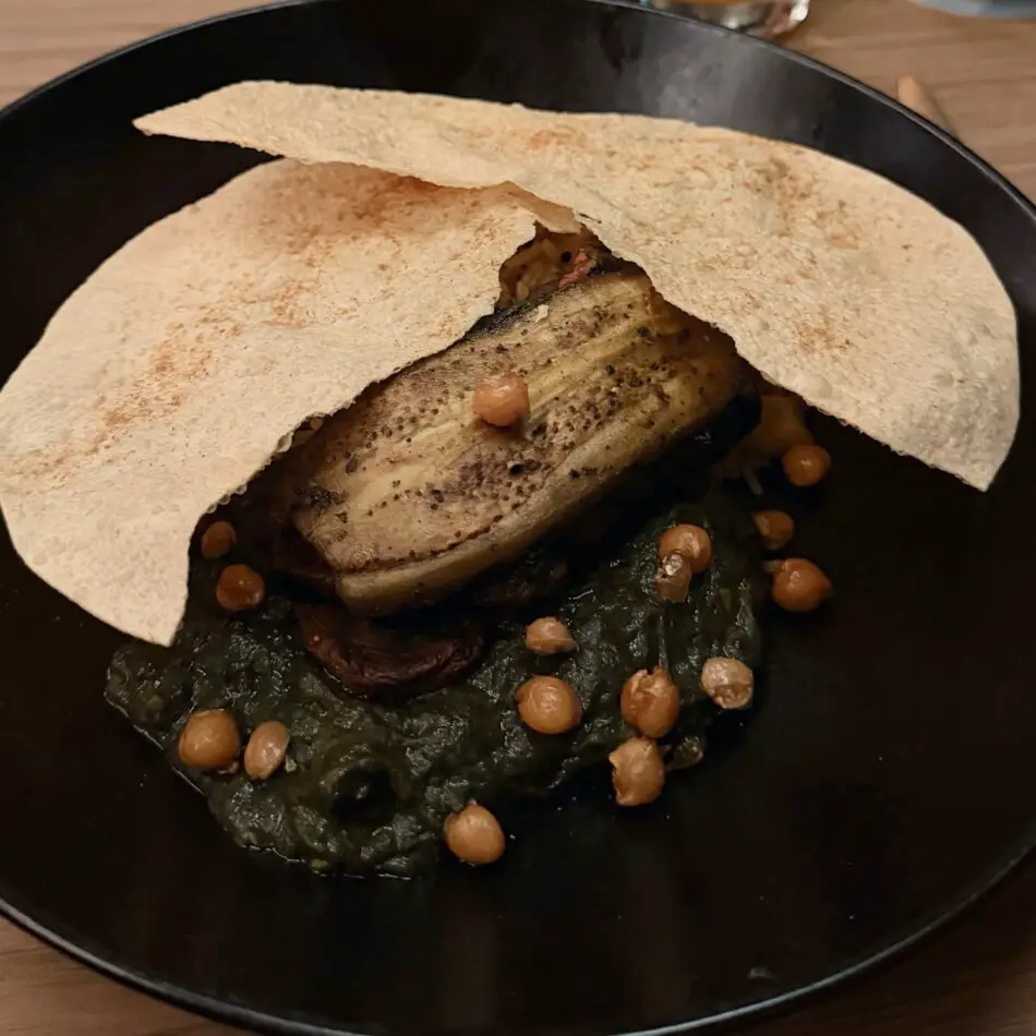 Seared eggplant on greens with chickpeas and flatbread on a dark plate
