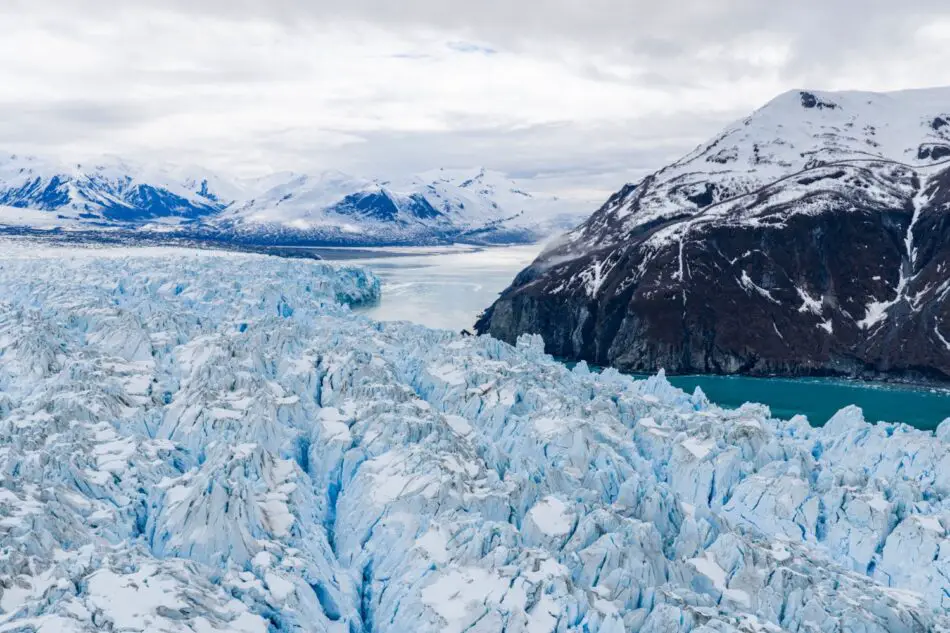 Blue glacier with jagged ice beside snow-covered mountains
