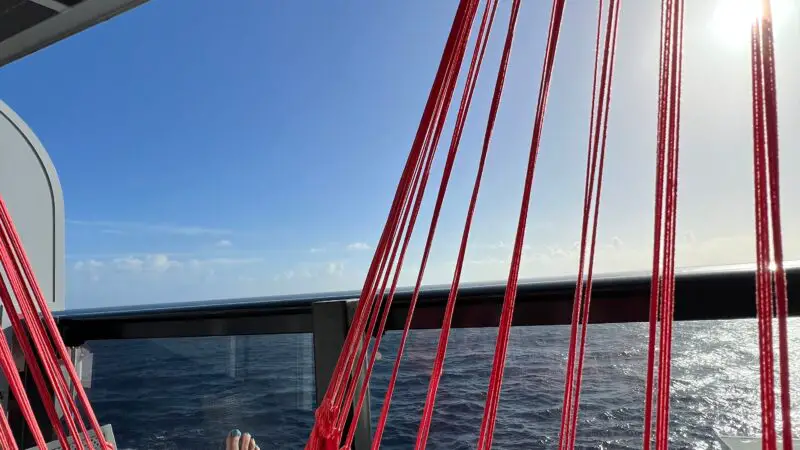 Person relaxing in a red hammock on a Virgin Voyages cruise ship balcony, with an ocean view and clear sky.