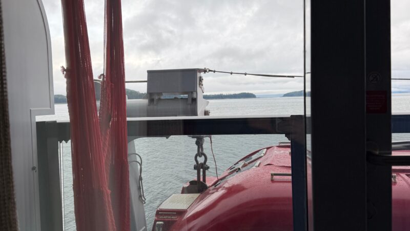 View from a Virgin Voyages cruise ship balcony cabin, featuring a red lifeboat, a small round table, and a red hammock against a seascape background.