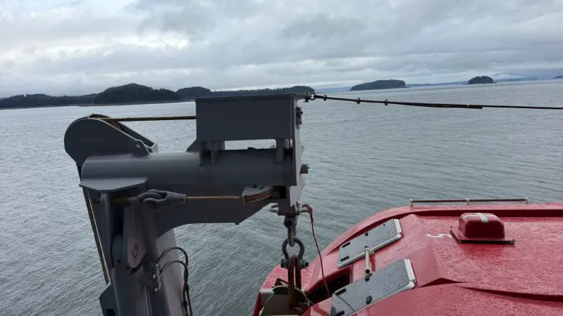 View from a Virgin Voyages cruise ship stateroom balcony, showing a lifeboat and calm sea under a cloudy sky.