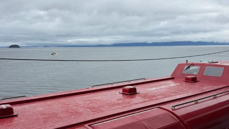 View from a Virgin Voyages cruise ship cabin balcony, showing the ocean, a red lifeboat below, and an overcast sky.