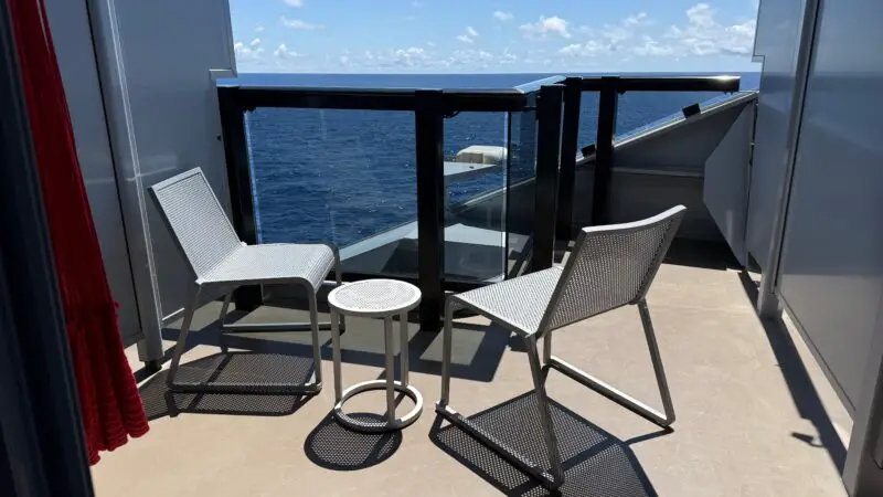 Balcony of a Virgin Voyages cruise ship cabin with two modern chairs and a small table, overlooking the ocean under a clear sky.
