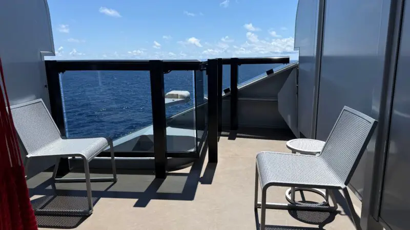Cruise ship cabin balcony with two white chairs, a small table, and an ocean view under a clear blue sky.