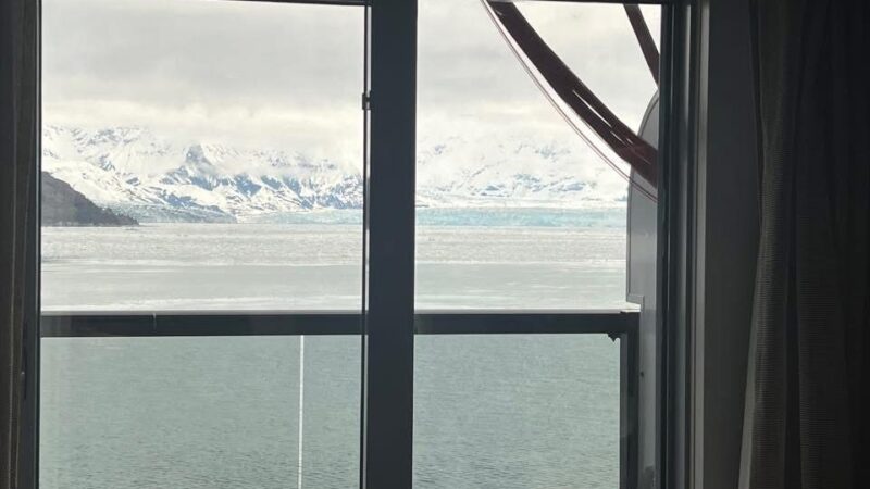 View from a Virgin Voyages cruise ship cabin showing a balcony with chairs and a snowy mountain landscape across the water.