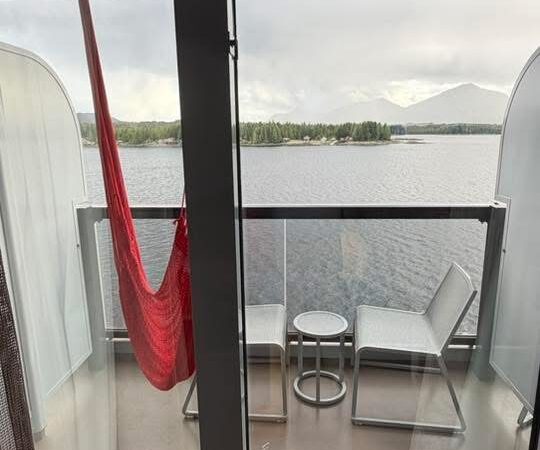 View from a Virgin Voyages cruise ship cabin showing a private balcony with two chairs, a small table, and a red hammock, overlooking the water and shoreline in the distance.