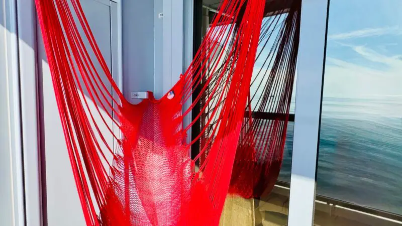 Red hammock on a balcony of a Virgin Voyages cruise ship cabin, with a glass door reflecting the ocean view.