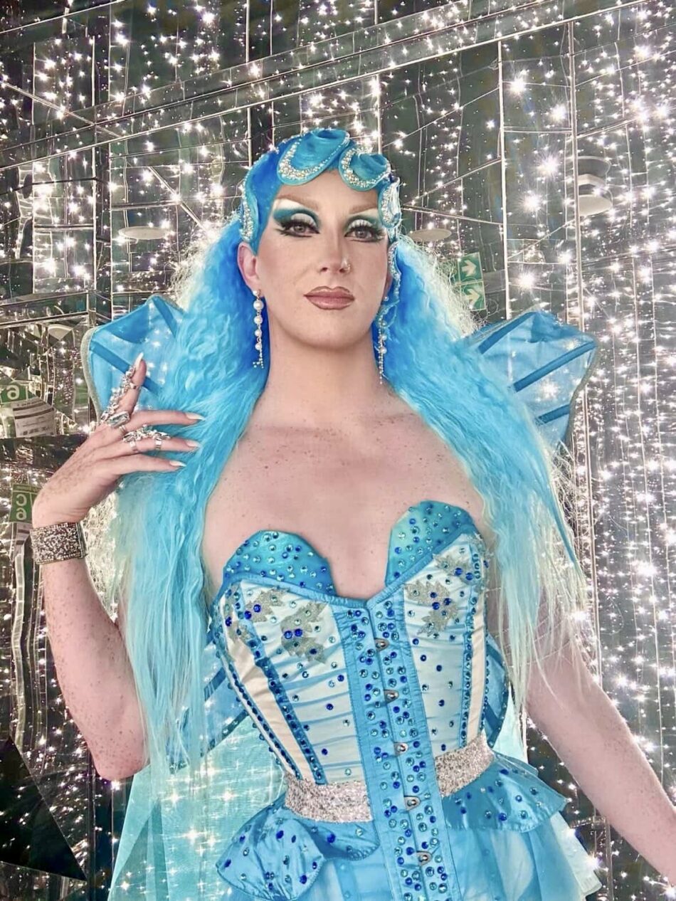 Drag performer in a vibrant blue sequined gown and turquoise wig posing among twinkling lights